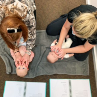 This image is taken from a bird's eye view and shows a 1 on 1 Baby Massage class. Mum is massaging babies lips as being instructed by our practitioner who is demonstrating on a toy baby.