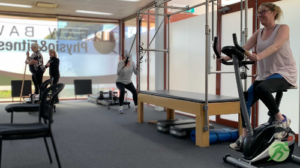 This image shows a Cancer Rehab Class. There are 3 participants and a practitioner. 1 participant is on the exercise bike, one is using the Trapeze Table to assist with squats, and the other is on the reformer bed and is being assisted by the practitioner.