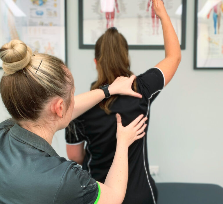 This image shows our Physiotherapist, Kate, assessing a clients shoulder range. The client has put their right arm up above their head. Kate has her hands on the upper and lower trap muscles to assess the range of movement.