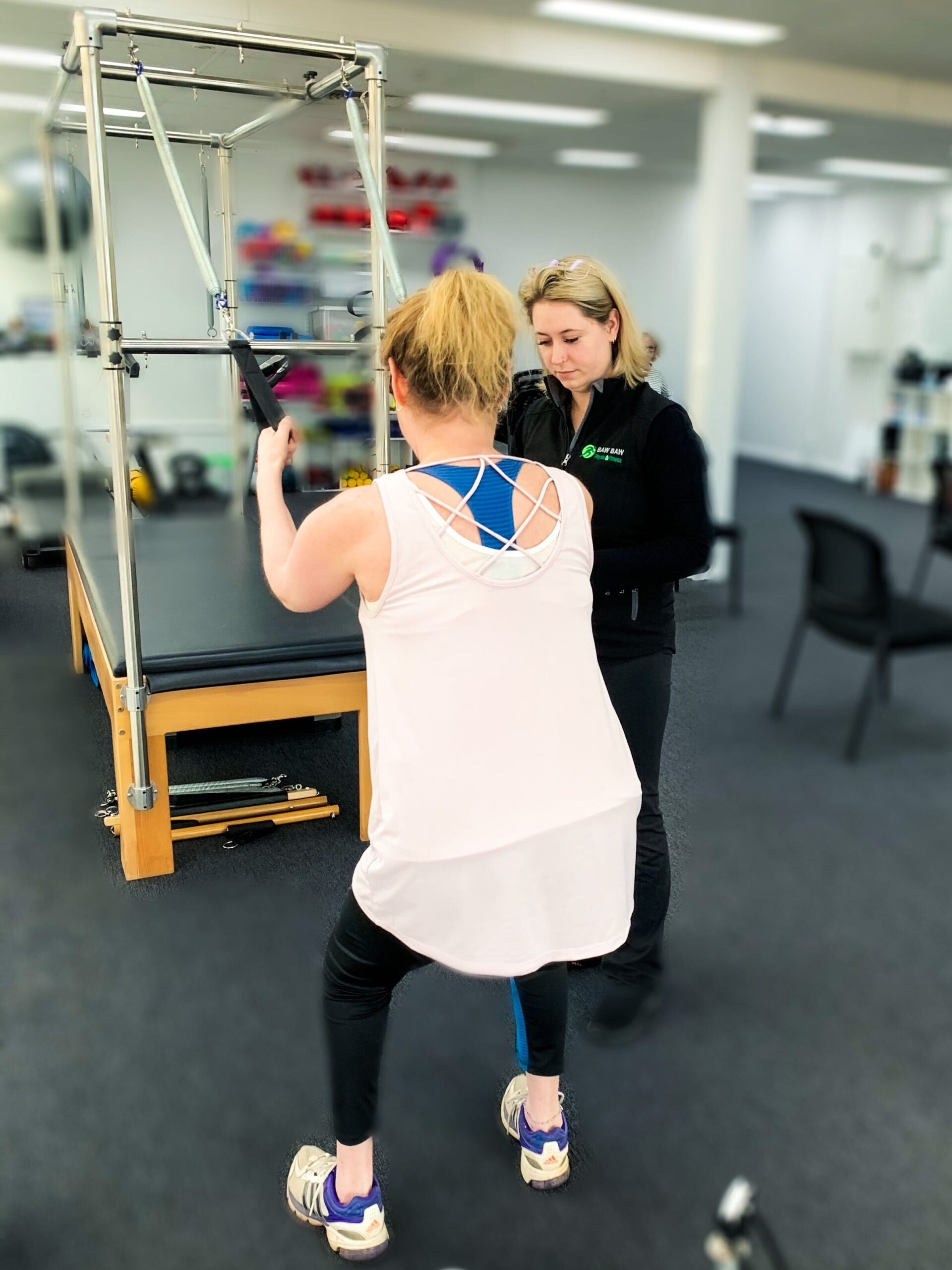 This image shows a client using our pilates trapeze table. The client is behind the trap table and using the hand pully's to assist them with a squat. A practitioner is supervising in the background.